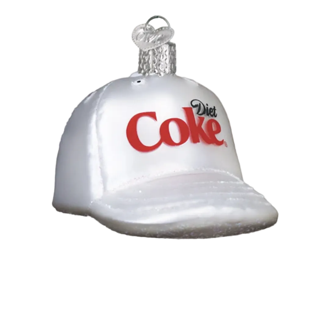 A silver Christmas ornament shaped like a baseball cap with the red and black Diet Coke logo, hanging from a tree branch with blurred holiday lights in the background.
