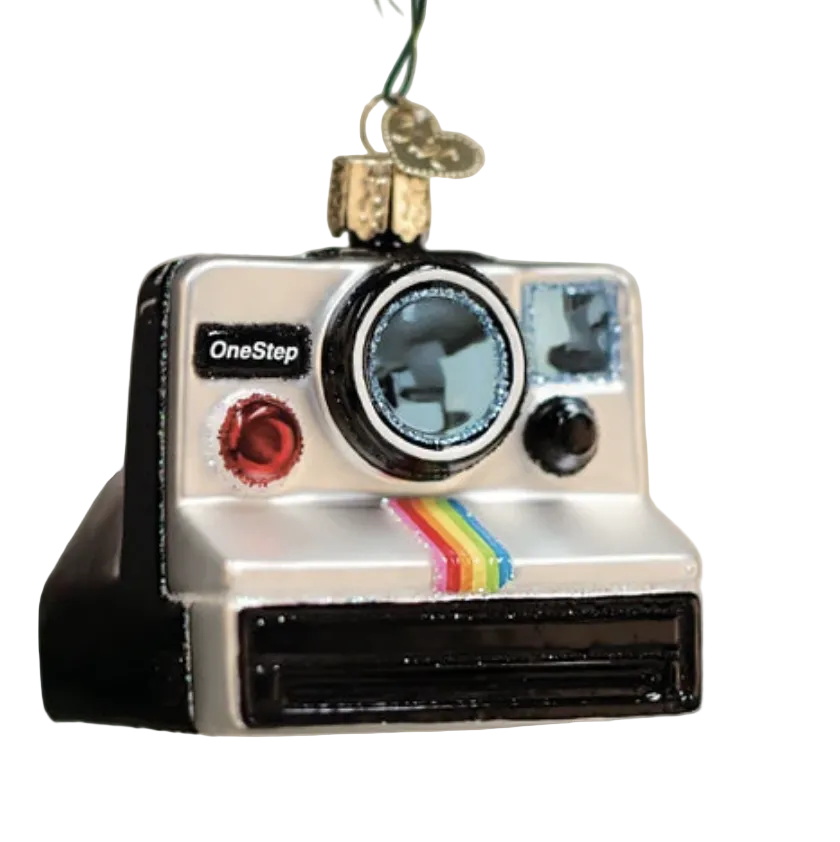 A Polaroid OneStep camera-shaped Christmas ornament hanging from a gold loop, with a small gift box attached above it. The ornament is silver and black with a rainbow stripe and the OneStep logo, set against a softly blurred holiday background with lights and evergreen branches.