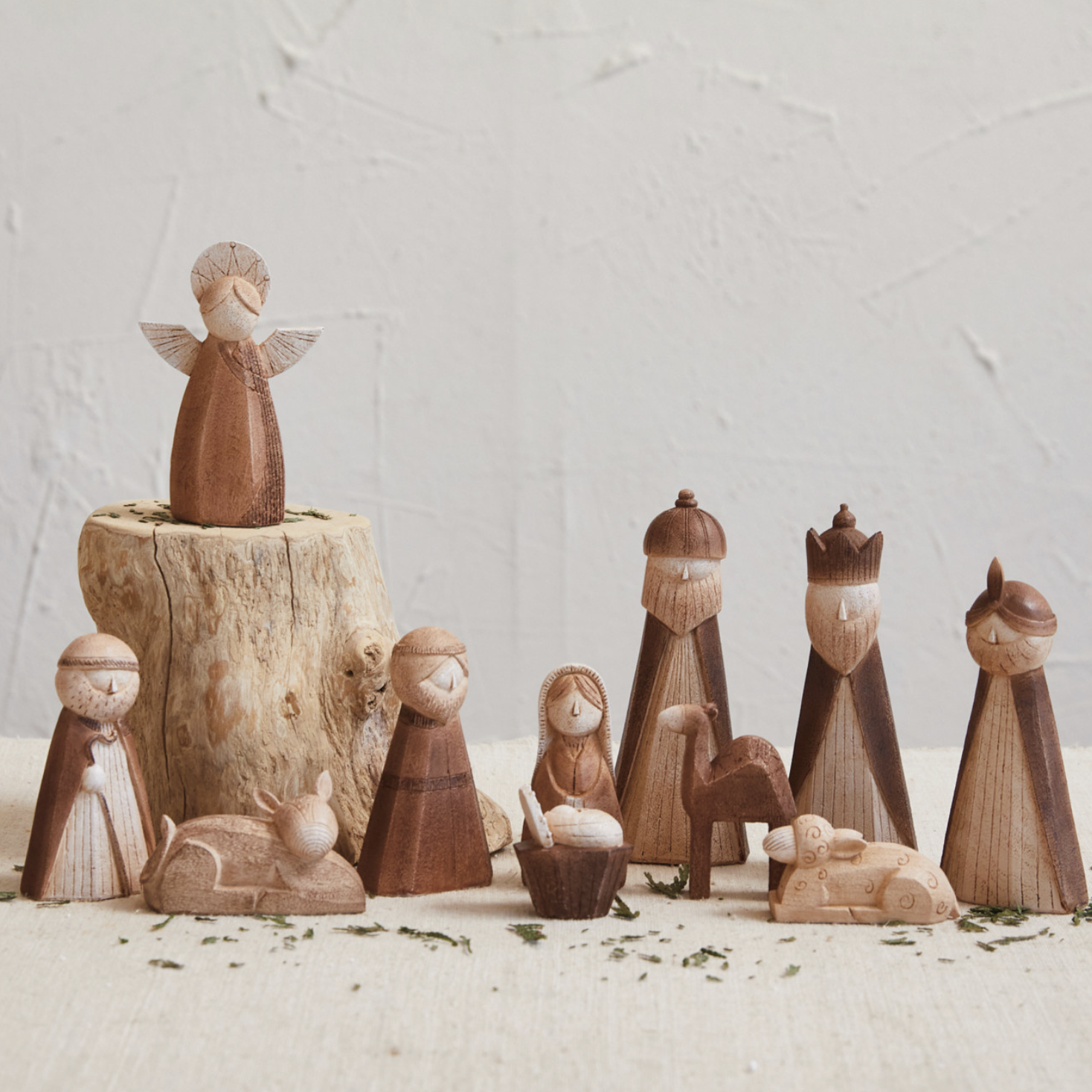 A set of hand-carved wooden nativity figurines arranged on a light surface against a textured off-white wall. The set includes an angel standing on a small tree stump, Mary, Joseph, baby Jesus in a manger, a camel, and three wise men.