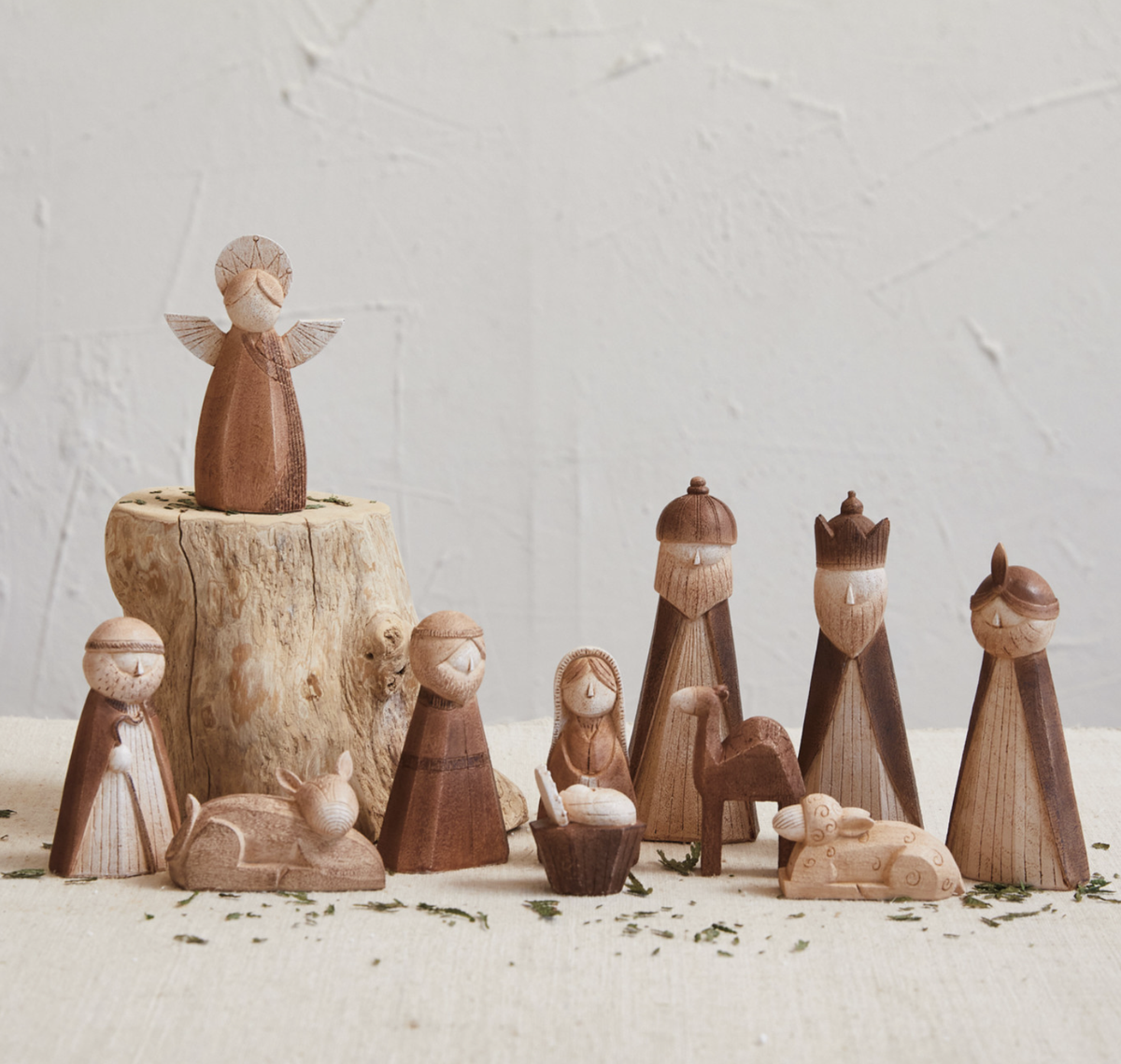 A set of hand-carved wooden nativity figurines arranged on a light surface against a textured off-white wall. The set includes an angel standing on a small tree stump, Mary, Joseph, baby Jesus in a manger, a camel, and three wise men.