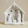 A white ceramic Christmas nativity scene figurine featuring Mary, Joseph, baby Jesus in a manger, and star-shaped cutouts, displayed on a textured beige surface against a neutral beige background.