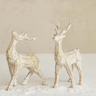 Two decorative reindeer figurines with a textured, weathered off-white finish, standing on a bed of white faux snow against a neutral beige background.
