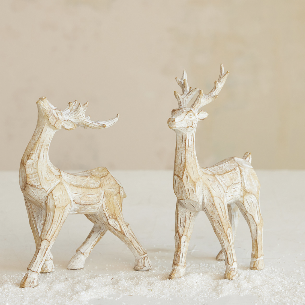 Two decorative reindeer figurines with a textured, weathered off-white finish, standing on a bed of white faux snow against a neutral beige background.
