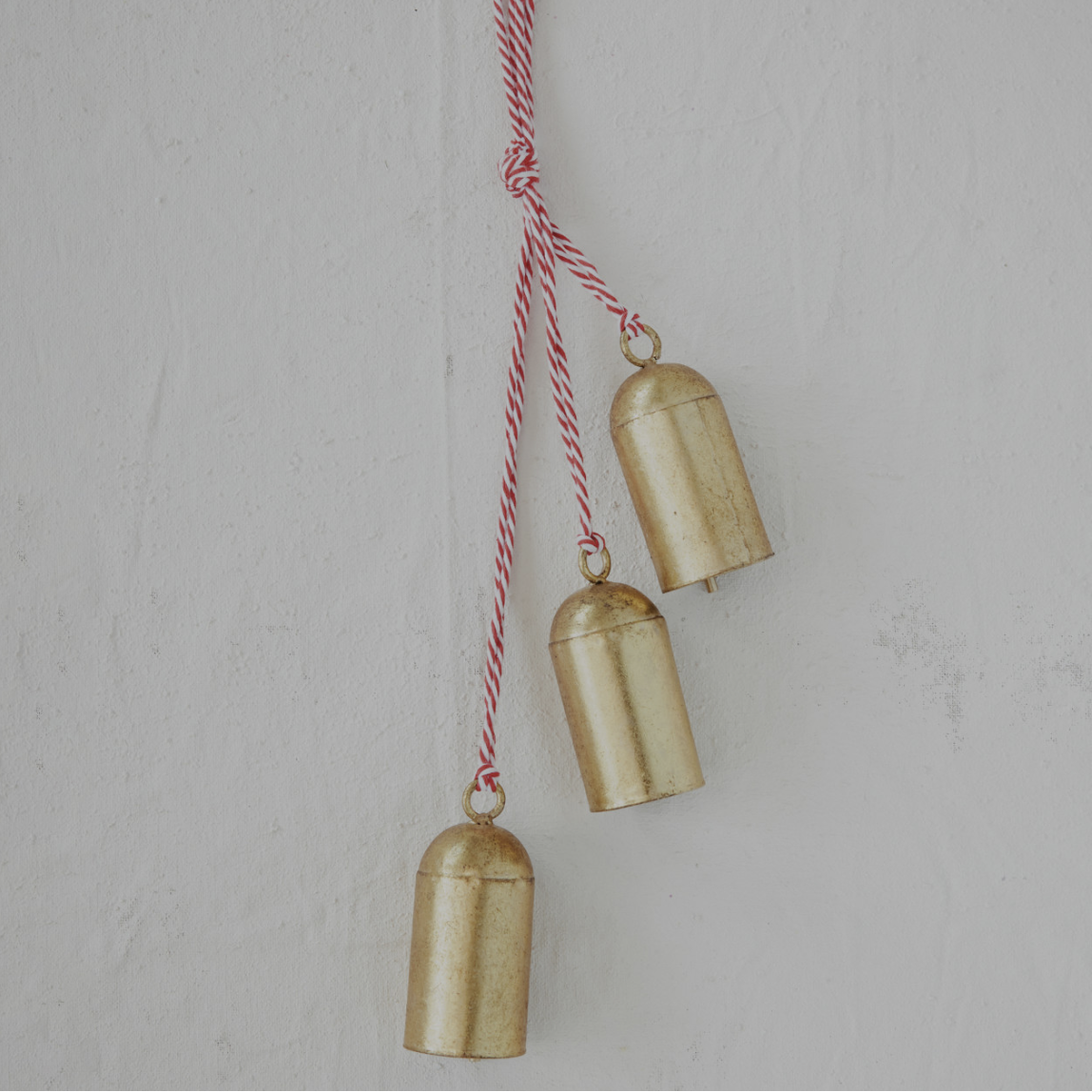 Three small gold bell-shaped hanging ornaments suspended from a red-and-white striped string against a light textured wall.
