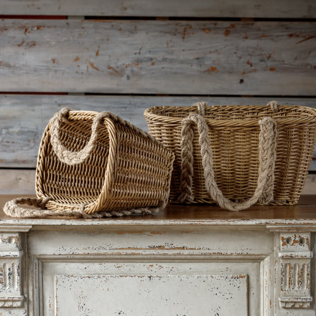 Two woven wicker baskets with thick rope handles, one standing upright and one lying on its side, displayed on a distressed white wooden cabinet against a rustic wood plank wall.