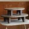 A rustic two-tier wooden coffee table with a dark weathered finish, set on a metal folding table against a warm wood-paneled background.