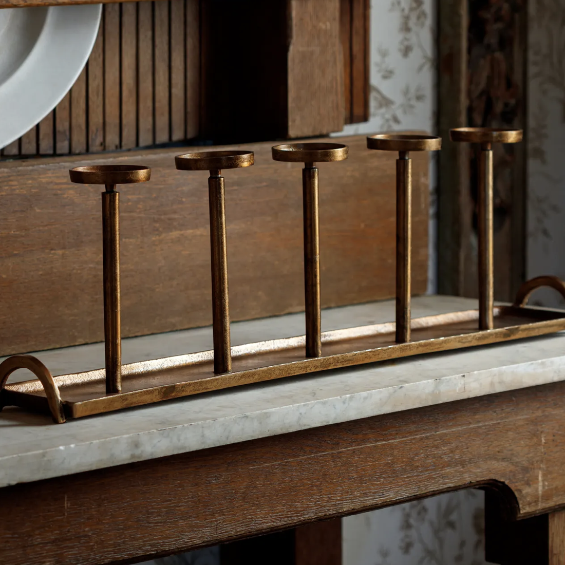 A brass five-stick candle holder with a rectangular base and arched ends, displayed on a marble countertop in a rustic kitchen setting.