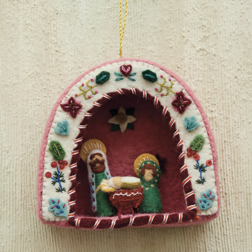 A handmade felt Christmas ornament shaped like a small arched nativity scene, hanging against a textured beige wall. The ornament features a pink border, a white embroidered outer frame with colorful floral motifs, and a dark red inner scene with small felt figures and a star.