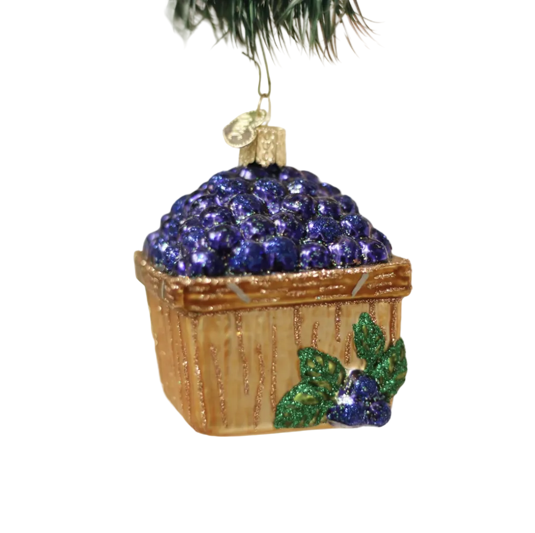 A glittery blueberry basket Christmas ornament hanging from a tree branch, with sparkling blueberries on top, a brown glittery basket base, and green leaves on the front, set against a soft blurred holiday background.