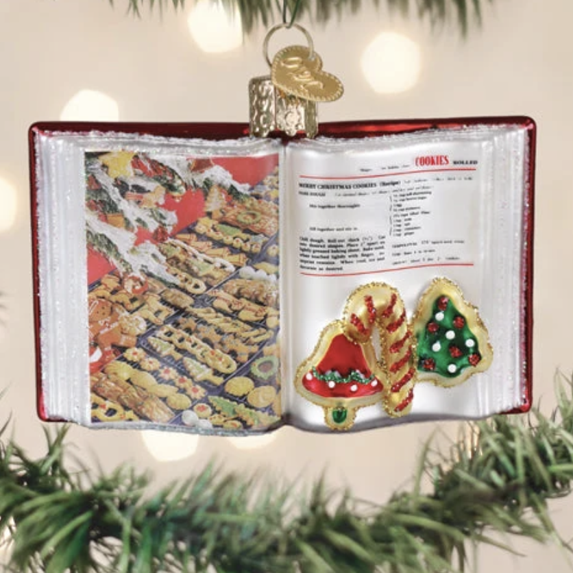 An open cookbook Christmas ornament hanging on a tree branch, with festive cookie cutouts on the pages and a gold hanger at the top.