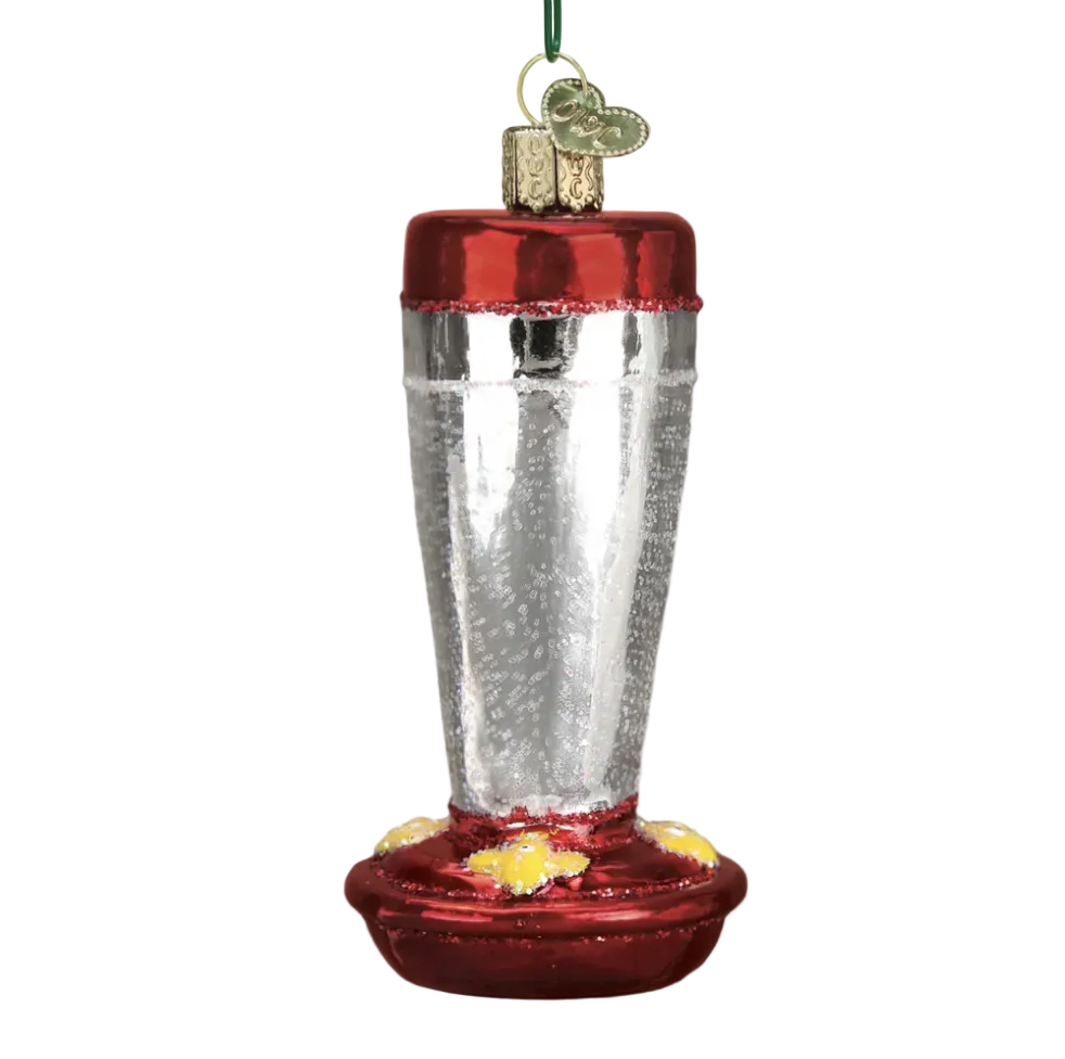 A Christmas ornament shaped like a bird feeder, with a textured silver body, a metallic red top and bottom, and small yellow accents near the feeder base. It hangs from a green string against a softly blurred background with warm bokeh lights and pine branches.