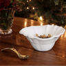 A white ceramic bowl with a scalloped rim sits on a wooden table, containing nuts. A small spoon rests nearby, with a blurred glass vase of flowers and warm holiday lights in the background.