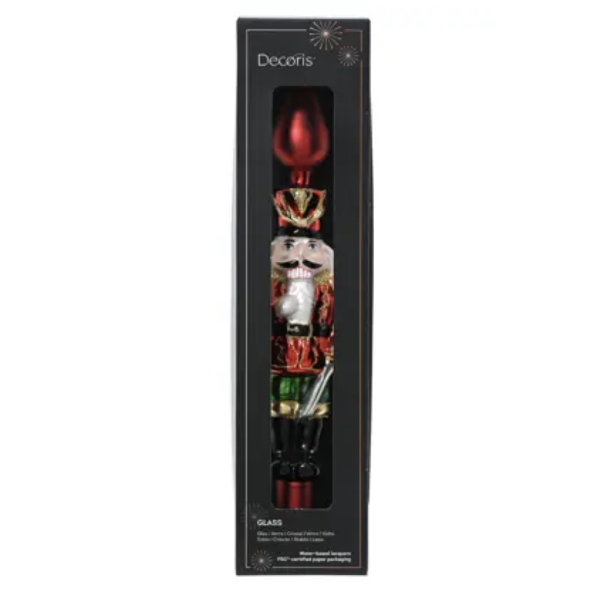 A decorative nutcracker-shaped glass candle in its original black retail packaging, featuring a red top and a detailed nutcracker figure with gold, red, white, and green accents.