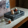 A dark metal egg tray with multiple circular egg holders sits on a light marble tabletop, with several decorative eggs placed inside and around it.