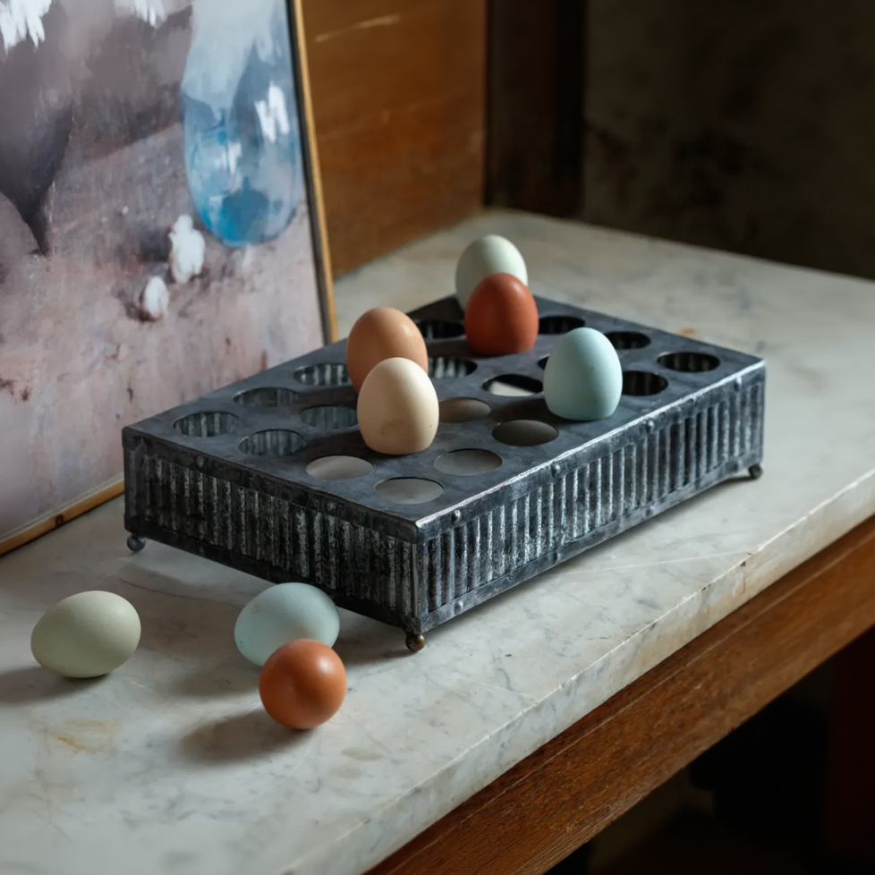 A dark metal egg tray with multiple circular egg holders sits on a light marble tabletop, with several decorative eggs placed inside and around it.