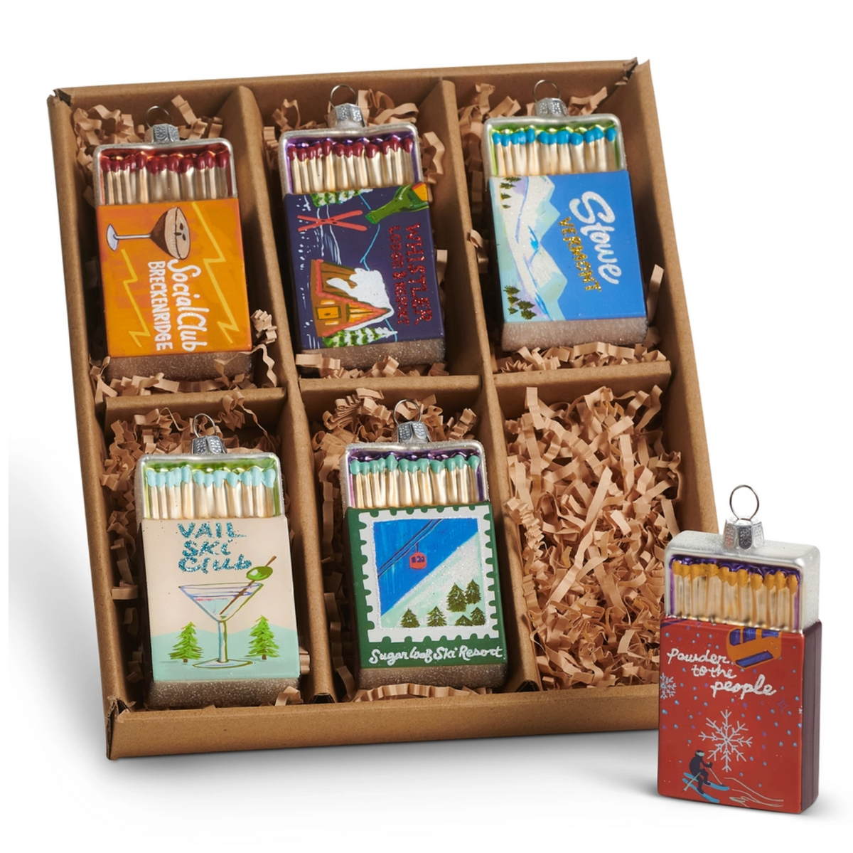 A boxed set of decorative matchbox ornaments displayed in a cardboard tray with shredded paper filler, plus one extra ornament standing beside the box on a white background.