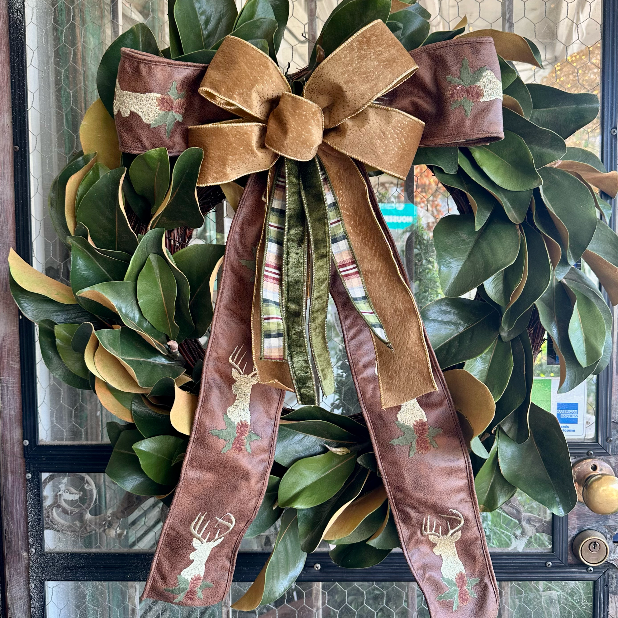 A festive holiday wreath made of green magnolia leaves with gold-tan edges, decorated with a large brown velvet bow and long ribbon tails featuring embroidered deer and holly motifs, hanging on a front door.