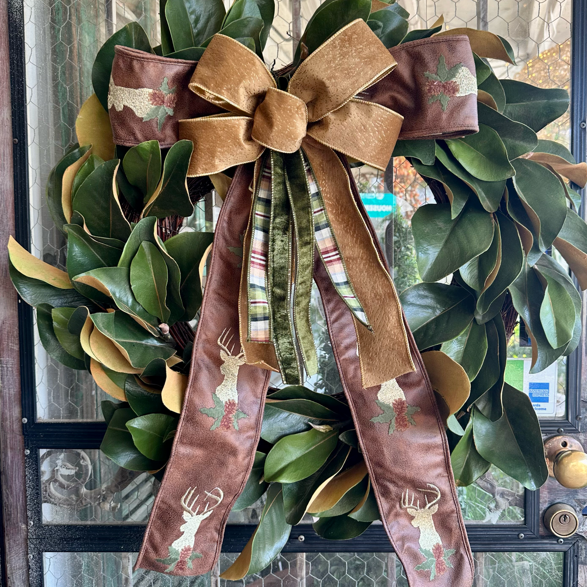 A festive holiday wreath made of green magnolia leaves with gold-tan edges, decorated with a large brown velvet bow and long ribbon tails featuring embroidered deer and holly motifs, hanging on a front door.
