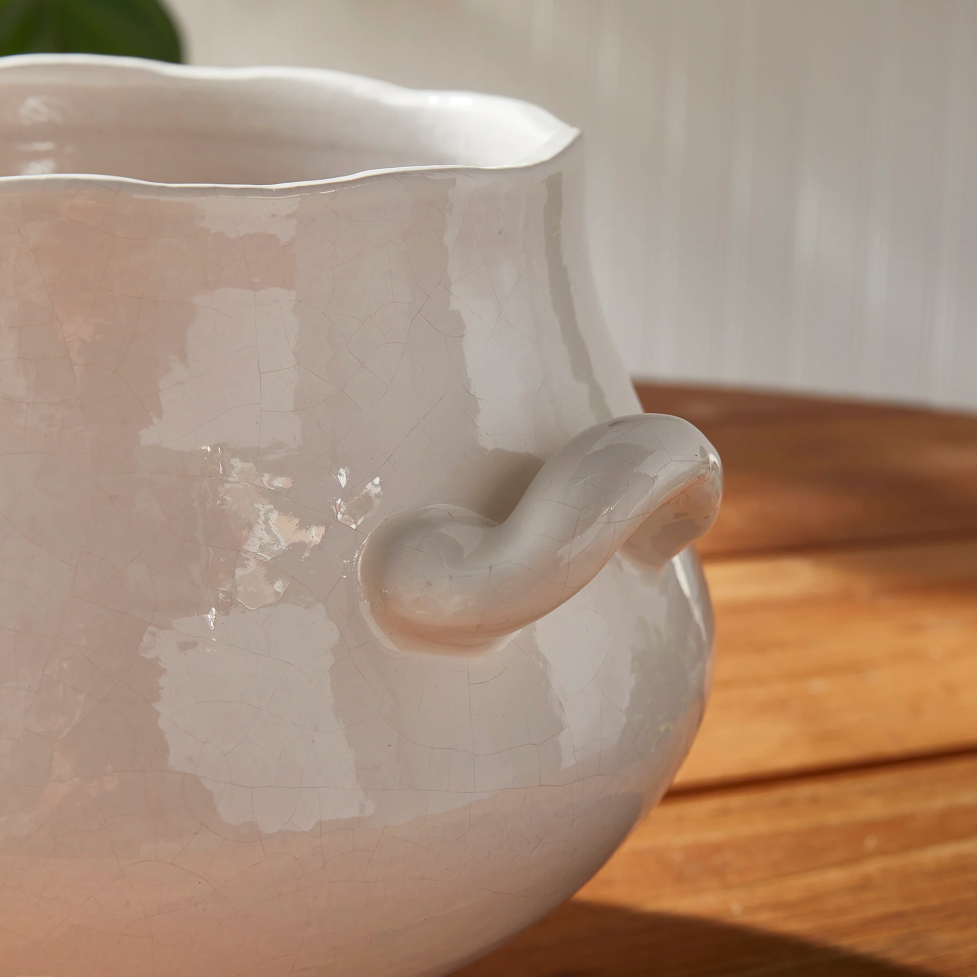 Close-up of a glossy off-white ceramic pitcher with a wavy rim and a curved side handle, resting on a wooden surface with soft natural light.
