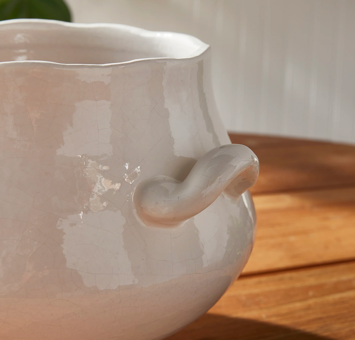 Close-up of a glossy off-white ceramic pitcher with a wavy rim and a curved side handle, resting on a wooden surface with soft natural light.