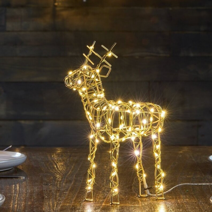 A golden wire-frame deer decoration illuminated with warm white string lights, displayed on a wooden table against a dark wood plank background.