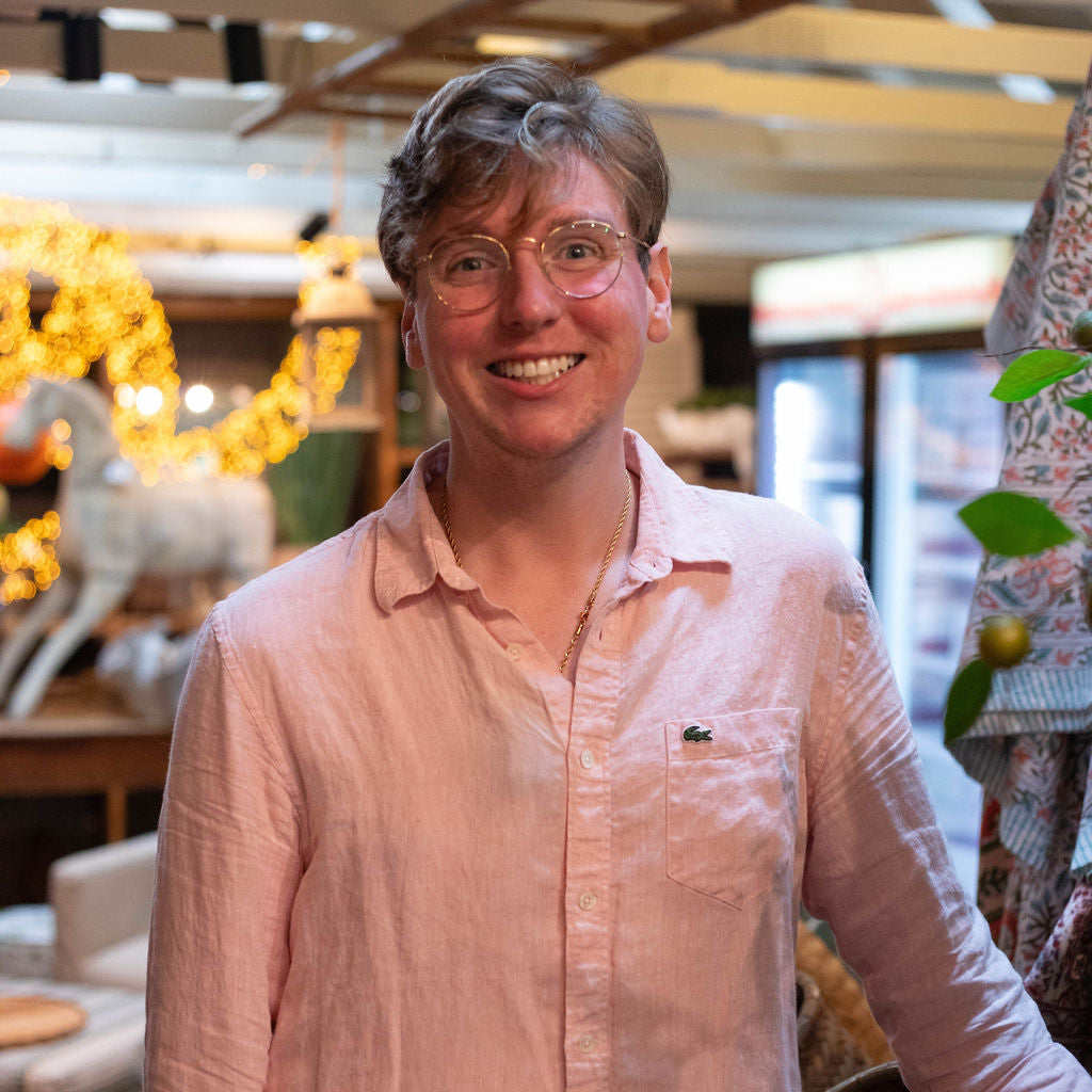 A smiling man with short light hair and glasses, wearing a light pink button-up shirt, standing indoors in a decorated retail or event space.