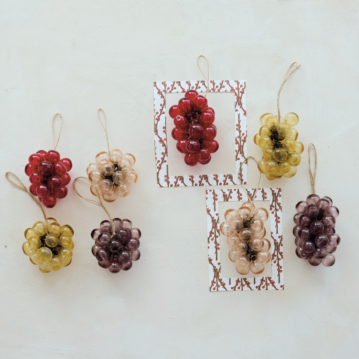 Six decorative hanging ornaments shaped like grape clusters in red, yellow, and purple tones, arranged on a light off-white background with two small patterned cards behind some of them.